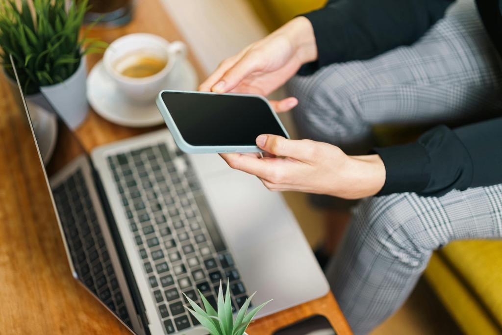 A person holds a smartphone above a laptop while reviewing a website experience at a desk.